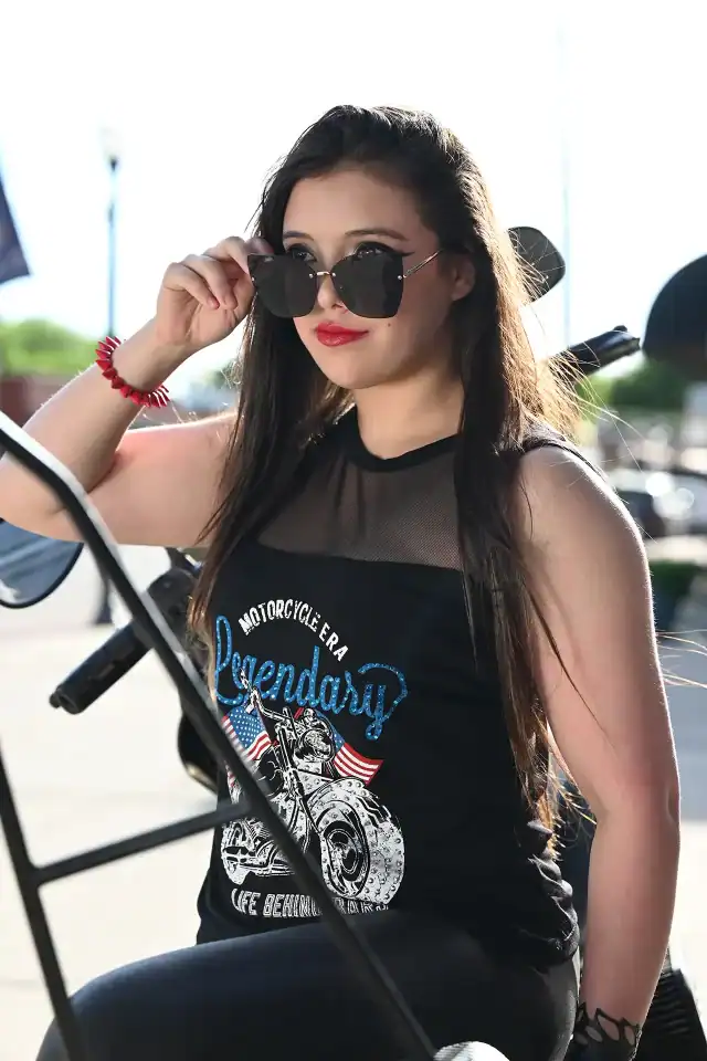 A woman wearing sunglasses and a black Liberty Wear tank top sitting on a motorcycle